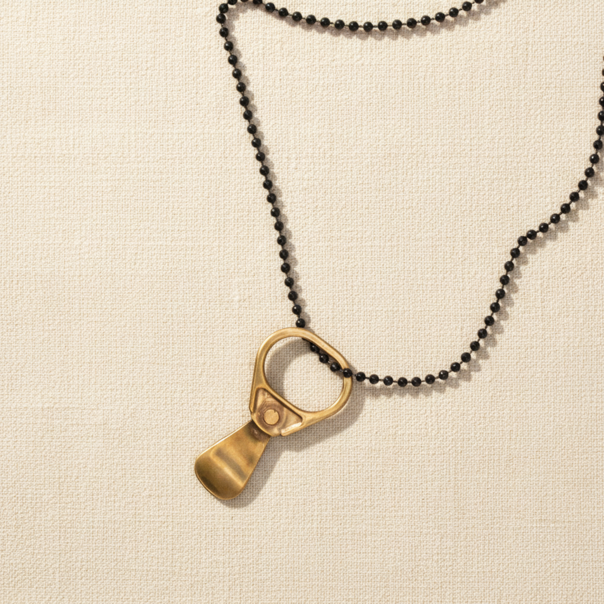 A black ball-chain necklace with a solid bronze pop top pendant on a linen background